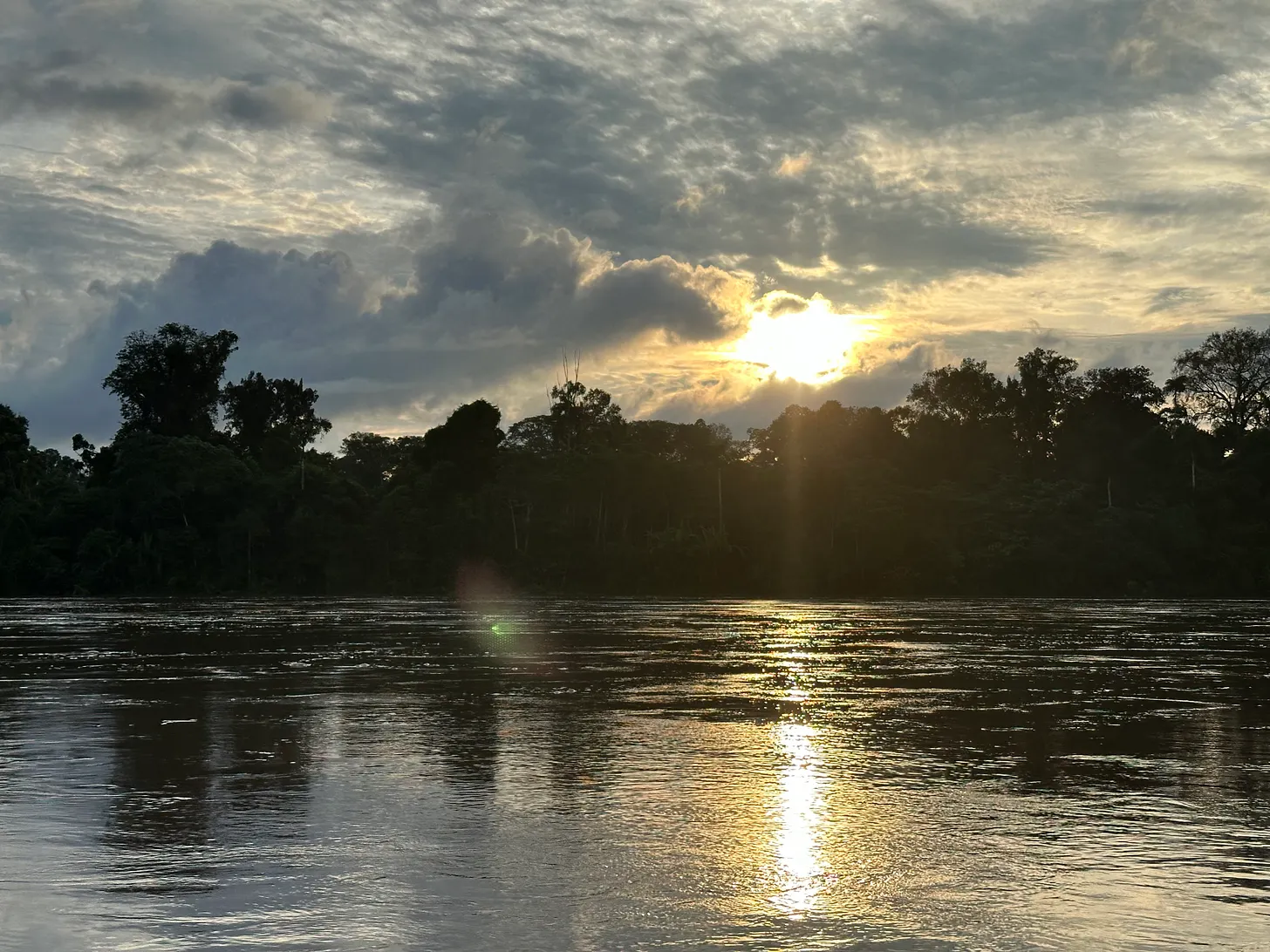A river at dusk, the sun breaking through cloud above the treeline.